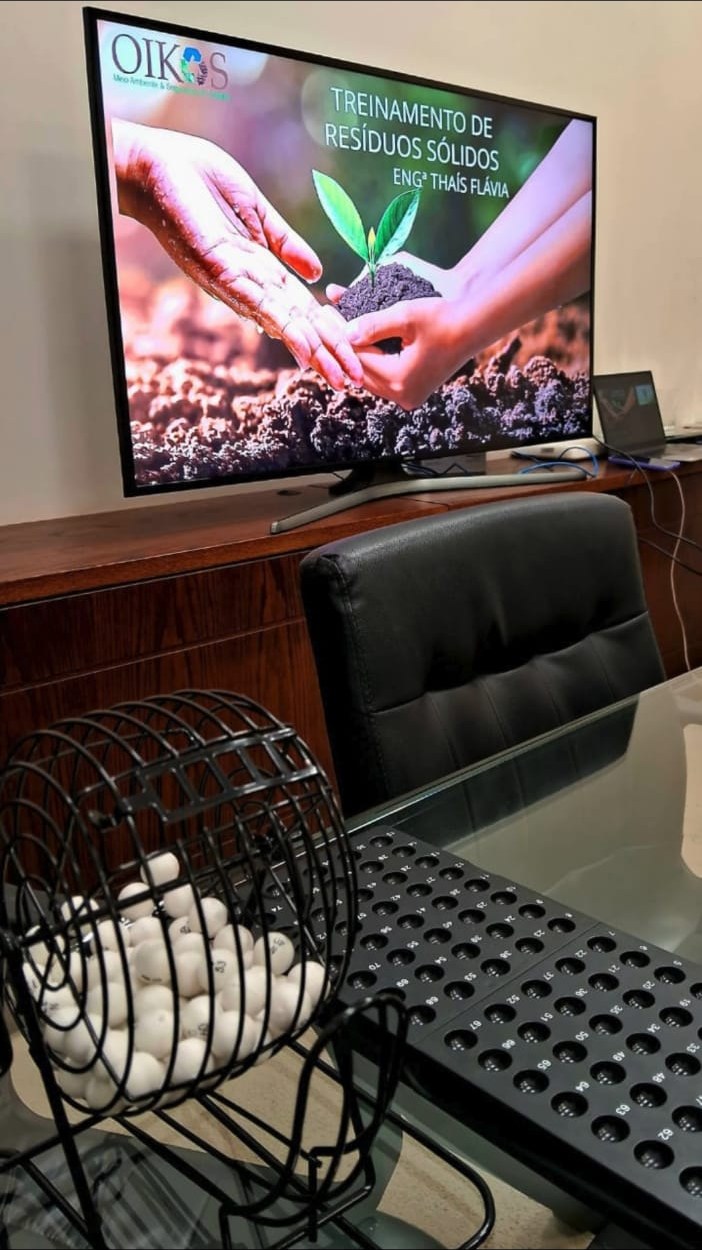 Bingo didático da OIKOS ao lado de uma TV com a apresentação do Treinamento de Resíduos Sólidos
